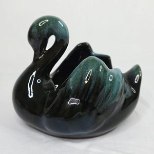 Puede incluir: Una maceta o cuenco decorativo de cerámica con forma de cisne y acabado brillante. La pieza es principalmente negra con reflejos en azul verdoso y verde oscuro en el cuello y las alas, creando una superficie lisa y muy reflectante.