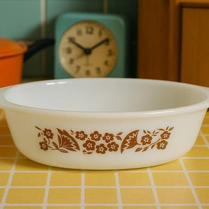 May include: White oval casserole dish with brown floral pattern. The dish has two handles and is set on a yellow tiled surface. An orange pot, a clock, and a wooden flour container are in the background.