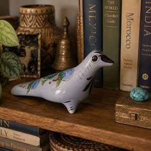May include: A light blue ceramic seal figurine featuring hand-painted floral motifs in green, blue, and brown, with black line details on the face and flippers. The smooth, glossy surface displays a stylized design with a pointed snout and eyes.