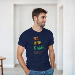 Könnte beinhalten: Marineblaues T-Shirt mit dem Text "EAT. SLEEP. GAME. REPEAT." in Orange, Grün und Grau. Das T-Shirt hat einen Rundhalsausschnitt und kurze Ärmel. Das Wort "GAME" enthält eine kleine Controller-Grafik.