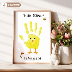 Puede incluir: Una impresión enmarcada con un pollito amarillo en forma de huella de mano, el texto "Frohe Ostern" e "Ich hab dich lieb", y dos abejas pequeñas. El fondo es blanco.