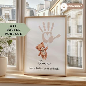 Puede incluir: Un cuadro enmarcado con una huella de mano y un corazón, un oso de peluche con globos y texto en alemán. El texto dice "Oma Ich hab dich ganz doll lieb". La obra está en un marco de madera.