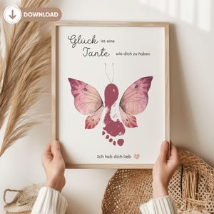 May include: A framed print featuring a butterfly design made from a footprint and watercolor wings in shades of pink and burgundy. The German text reads "Glück ist eine Tante wie dich zu haben" and "Ich hab dich lieb".