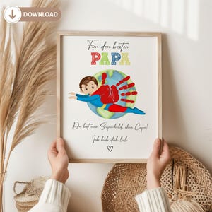 May include: A framed print with the words "Für den besten PAPA" and a drawing of a superhero with a handprint cape. The print includes the phrases "Du bist mein Superheld, ohne Cape!" and "Ich hab dich lieb" with a heart symbol.
