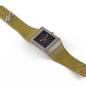 May include: A square-shaped wristwatch with a black face and a green leather band. The watch has a unique design with a red hand and a black and white pattern on the face.