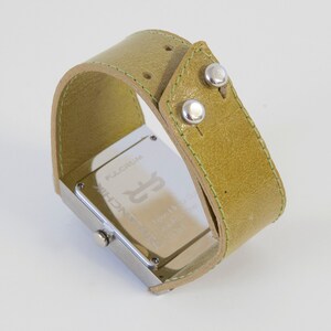 May include: A silver watch with a square face and a green leather band. The watch face has the words "FULLCRUM" and "TECHNIC" printed on it.