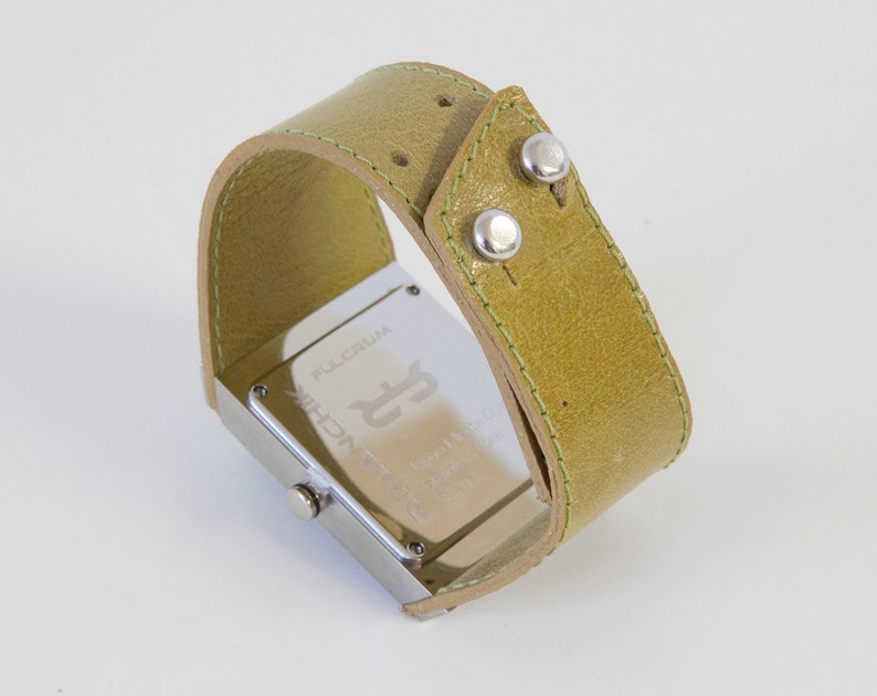 May include: A silver watch with a square face and a green leather band. The watch face has the words "FULLCRUM" and "TECHNIC" printed on it.