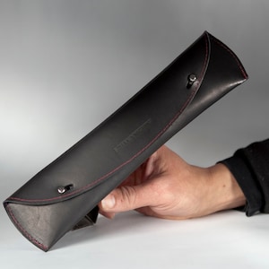 May include: Black leather pencil case with red stitching and silver snaps. The case is rolled up and held in a hand.