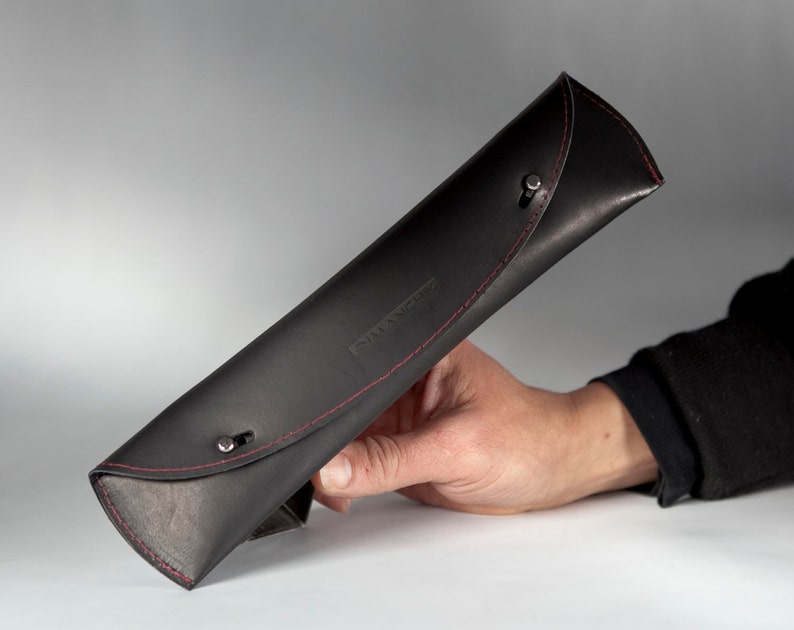 May include: Black leather pencil case with red stitching and silver snaps. The case is rolled up and held in a hand.