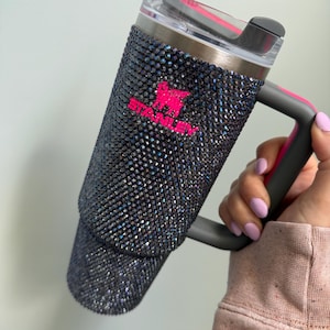 Pu&ograve; includere: Un bicchiere Stanley nero tempestato di strass iridescenti, con logo e dettagli del manico rosa. Il bicchiere ha un coperchio trasparente e una cannuccia riutilizzabile. Il manico &egrave; grigio con un accento rosa.