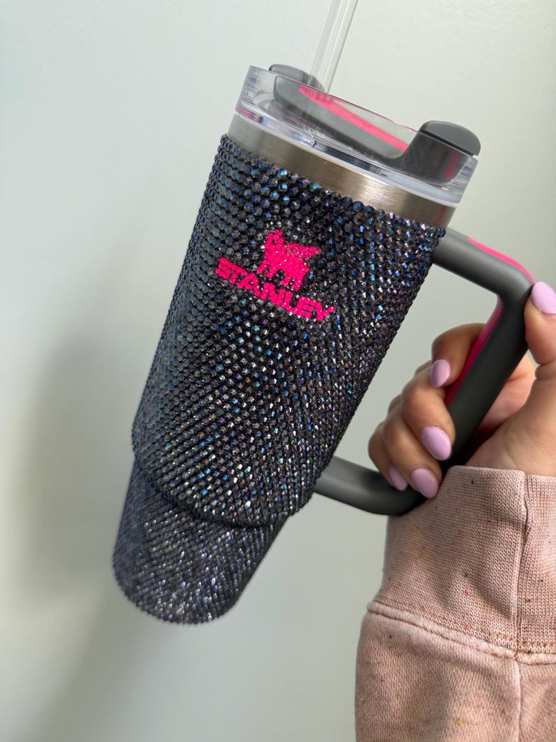Pu&ograve; includere: Un bicchiere Stanley nero tempestato di strass iridescenti, con logo e dettagli del manico rosa. Il bicchiere ha un coperchio trasparente e una cannuccia riutilizzabile. Il manico &egrave; grigio con un accento rosa.