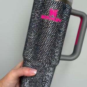 Pu&ograve; includere: Un grande bicchiere isolante ricoperto di scintillanti strass iridescenti. Il bicchiere ha un manico grigio con un accento rosa e un coperchio grigio. Il logo rosa Stanley &egrave; visibile sul davanti.