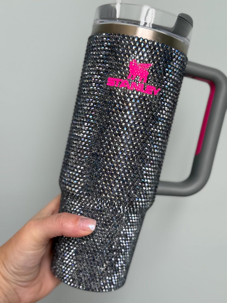 Pu&ograve; includere: Un grande bicchiere isolante ricoperto di scintillanti strass iridescenti. Il bicchiere ha un manico grigio con un accento rosa e un coperchio grigio. Il logo rosa Stanley &egrave; visibile sul davanti.