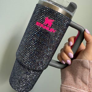 Pu&ograve; includere: Un grande bicchiere isolato con un esterno nero e glitterato. Il bicchiere ha un manico e un coperchio grigi, con un logo rosa. Una cannuccia trasparente &egrave; inserita nel coperchio. Il bicchiere &egrave; progettato per mantenere le bevande fredde o calde.