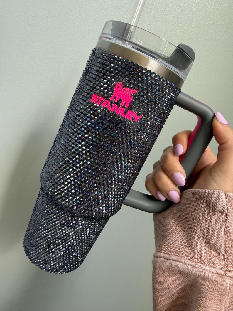 Pu&ograve; includere: Un grande bicchiere isolato con un esterno nero e glitterato. Il bicchiere ha un manico e un coperchio grigi, con un logo rosa. Una cannuccia trasparente &egrave; inserita nel coperchio. Il bicchiere &egrave; progettato per mantenere le bevande fredde o calde.