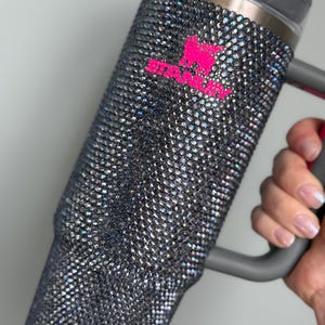 Pu&ograve; includere: Un bicchiere tempestato di strass con manico e coperchio grigi. Il bicchiere &egrave; ricoperto di piccoli strass iridescenti. Un logo rosa brillante &egrave; visibile sul lato del bicchiere.