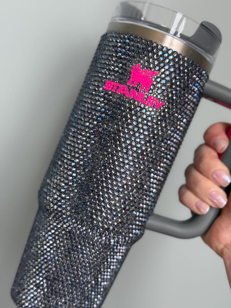 Pu&ograve; includere: Un bicchiere tempestato di strass con manico e coperchio grigi. Il bicchiere &egrave; ricoperto di piccoli strass iridescenti. Un logo rosa brillante &egrave; visibile sul lato del bicchiere.