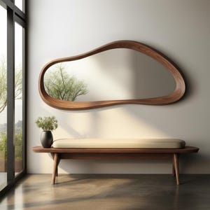 May include: A modern, organic-shaped wooden mirror hangs above a wooden bench with a cream-colored cushion. A black vase with greenery sits on the bench. The mirror reflects a tree and the room's natural light.