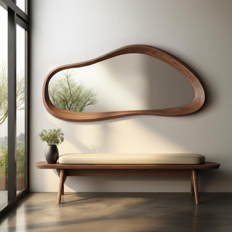 May include: A modern, organic-shaped wooden mirror hangs above a wooden bench with a cream-colored cushion. A black vase with greenery sits on the bench. The mirror reflects a tree and the room's natural light.