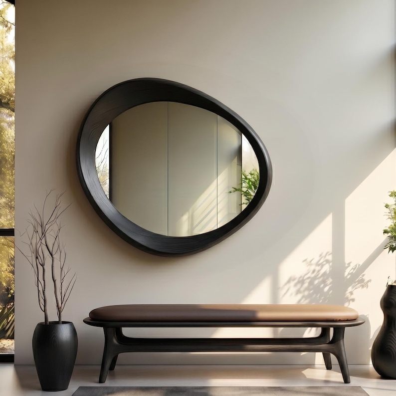 May include: A wall-mounted mirror with an irregular, rounded black frame. Below the mirror sits a long, low bench featuring a dark wooden base and a smooth, brown upholstered seat. The mirror reflects a bright interior space with light walls.