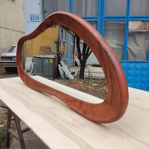 May include: A large, organically shaped mirror with a rich, reddish-brown wooden frame. The mirror is positioned horizontally on a light-colored wooden surface, reflecting the outdoor environment. The frame has a smooth, curved design.