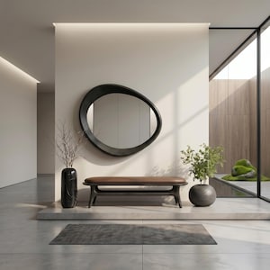 May include: An oval, dark-framed mirror hangs above a low bench with a brown seat and black base. To the left, a tall black vase holds bare branches, while a round black pot containing a green plant sits on the right side of the bench.
