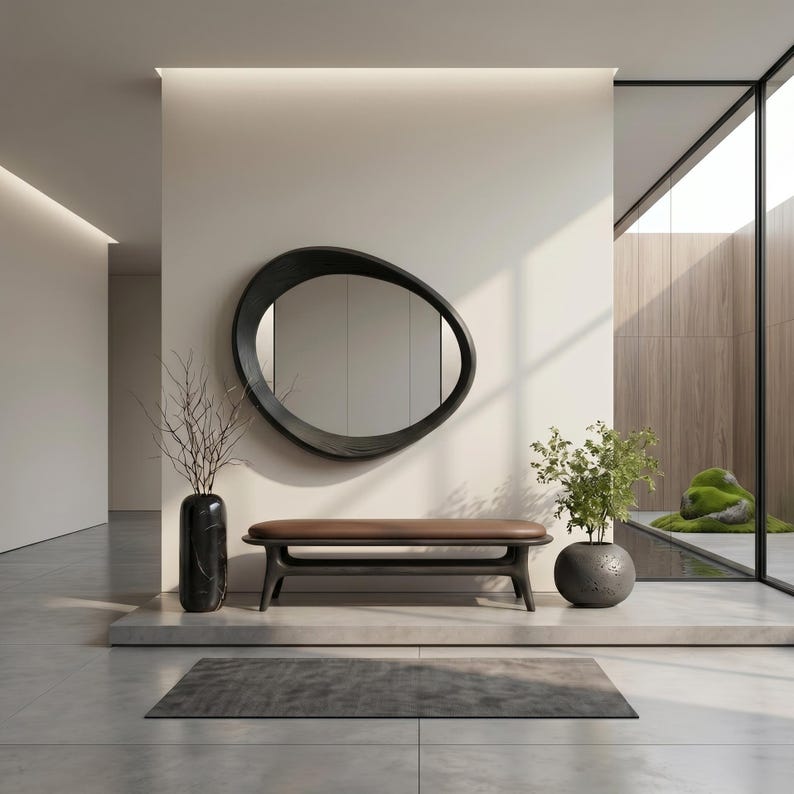 May include: An oval, dark-framed mirror hangs above a low bench with a brown seat and black base. To the left, a tall black vase holds bare branches, while a round black pot containing a green plant sits on the right side of the bench.