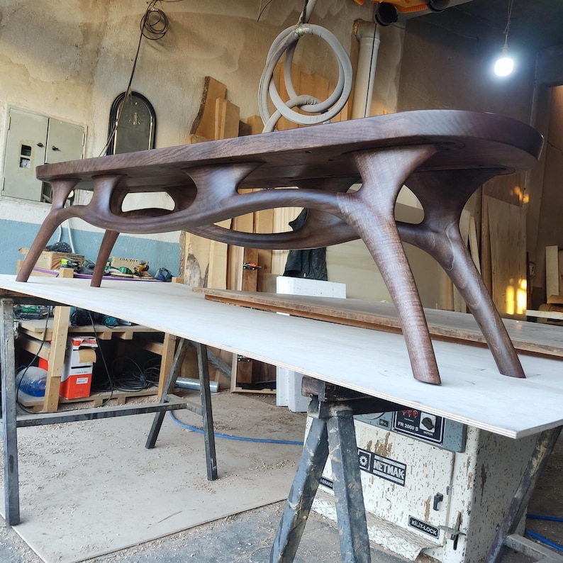 May include: A handcrafted, dark brown wooden table with a unique, organic design. The table features a smooth, oval-shaped top and intricately carved legs, showcasing a modern aesthetic. The table's dimensions are approximately 72 inches long, 30 inches wide, and 18 inches high.
