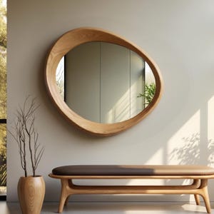 May include: An asymmetrical, organic-shaped wall mirror with a light-toned, natural wood grain frame. The mirror reflects a bright interior space, showcasing a smooth, curved wooden border that encircles the reflective glass surface.
