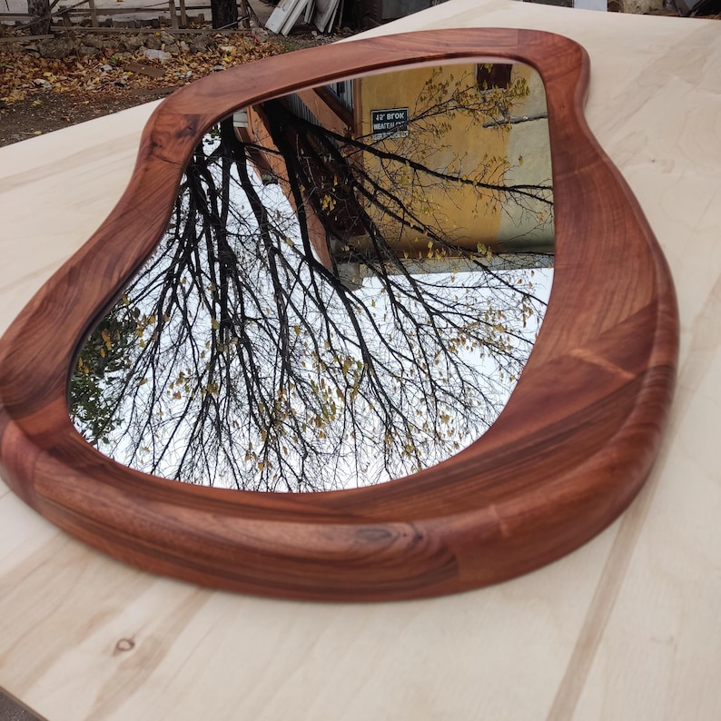 May include: A unique, free-form wooden mirror with a rich, reddish-brown frame. The mirror reflects a tree with yellow leaves and a building. The frame has a smooth, curved design, adding a natural and artistic touch to the decor.