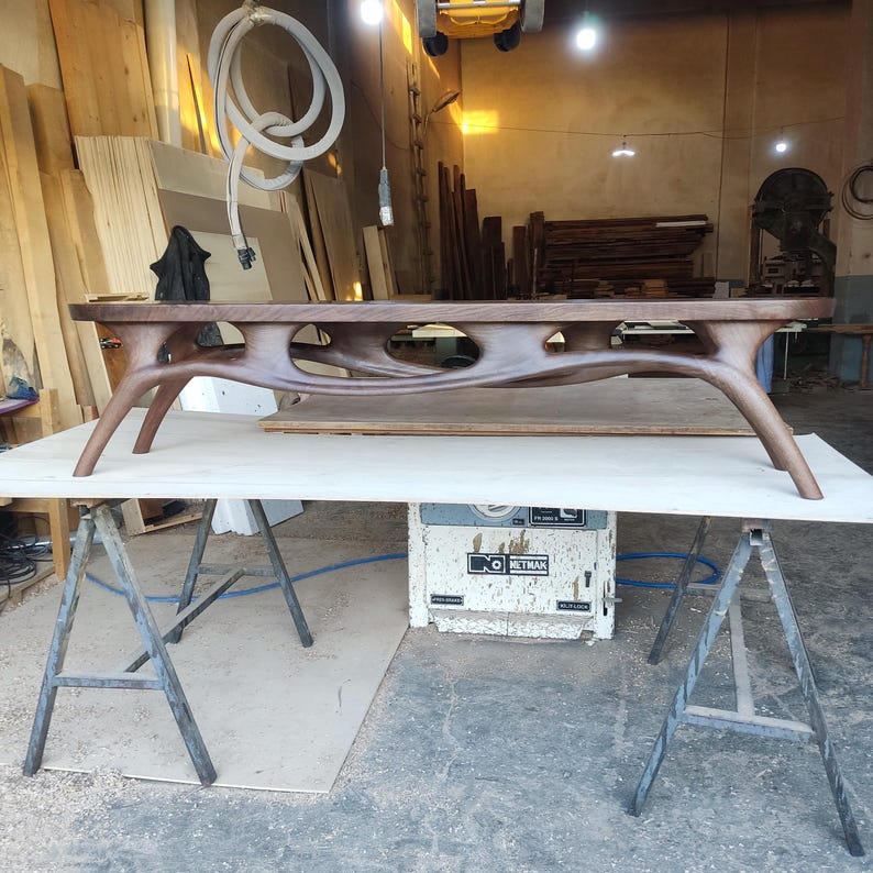May include: A handcrafted, dark brown wooden coffee table with a unique, organic design. The table features curved legs and a carved central support with open spaces. It is displayed on a light-colored surface in a workshop setting.