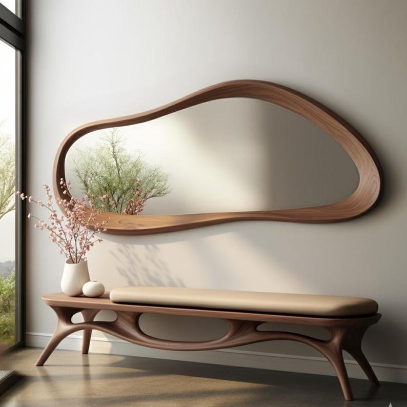 May include: A modern, organic-shaped wooden mirror with a shelf is mounted on a neutral wall. Below, a matching wooden bench with a beige cushion complements the mirror. The room is decorated with a vase of flowers.