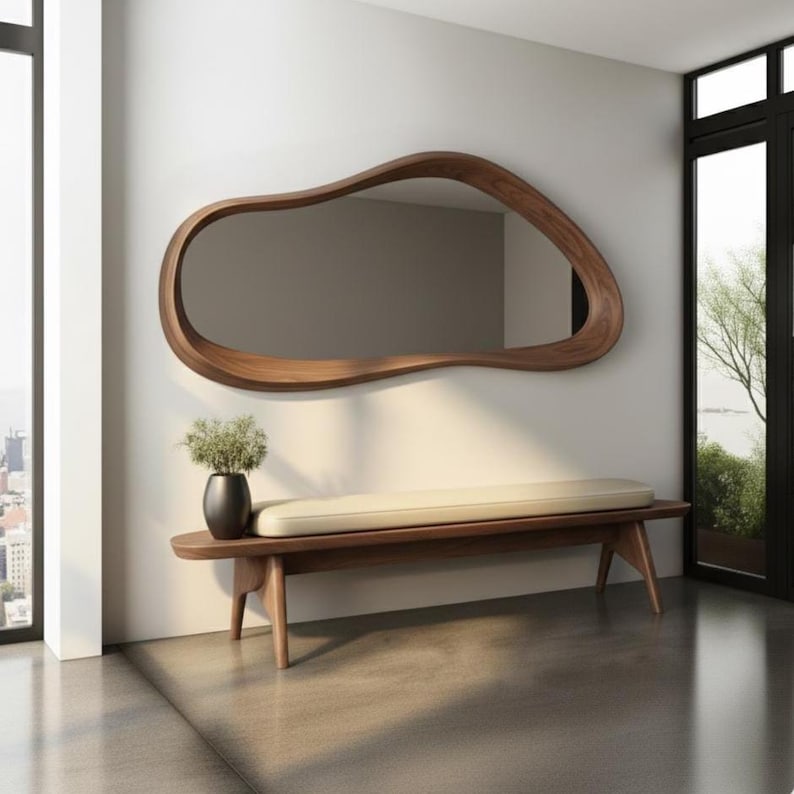 May include: A large, organically shaped wooden mirror hangs above a bench with a cream-colored cushion. The mirror's frame is a rich brown, and the bench has a matching wooden frame. A black vase with greenery sits on the bench.