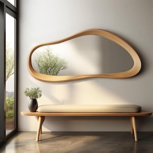 May include: A large, horizontal, organic-shaped wall mirror with a smooth, light-toned wooden frame. Below the mirror sits a long, rectangular wooden bench with a cushioned seat and a small black vase containing a green plant on one side.