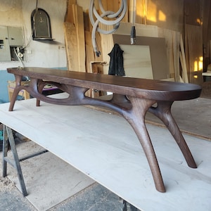 May include: A handcrafted, dark brown wooden bench with a unique, organic design. The bench features a long, curved seat supported by intricately carved legs. The bench is approximately 72 inches long.