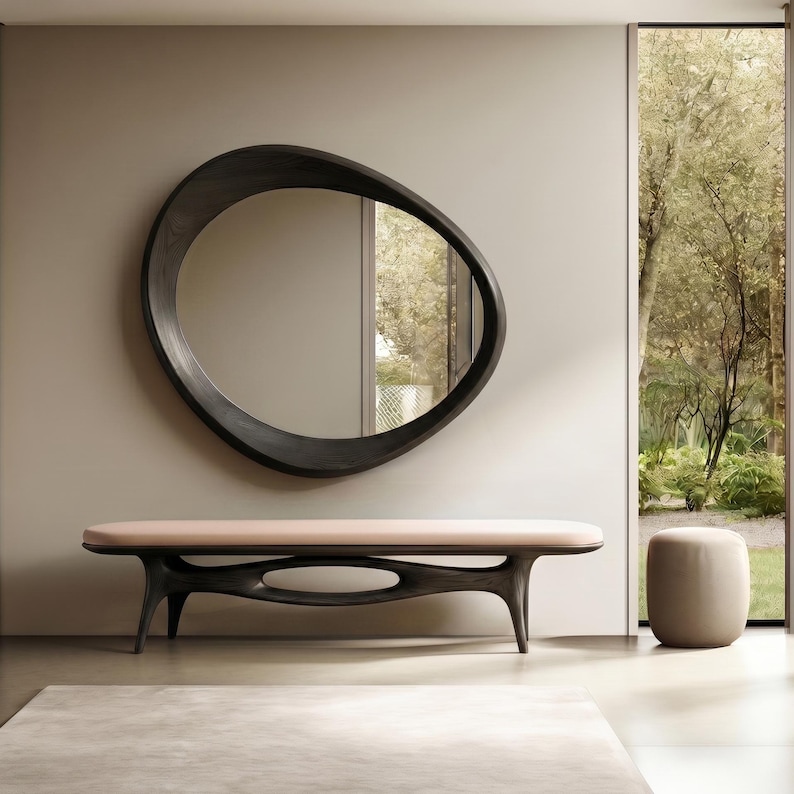 May include: A dark wood wall mirror with an irregular, organic, pebble-like shape. Below it sits a long bench with a light pink upholstered seat and a dark wood base featuring a central cutout, accompanied by a small, matching light pink stool.