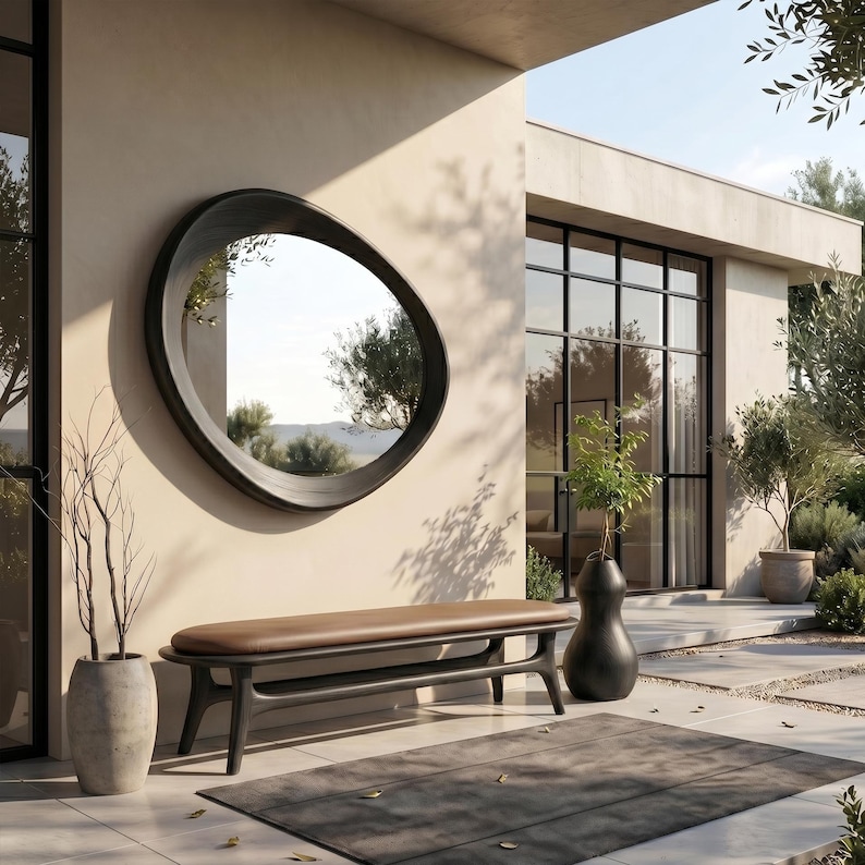 May include: A large, oval-shaped wall mirror with a thick, dark, rounded frame mounted on a beige wall above a low bench. The bench features a smooth, brown leather-like seat cushion supported by a dark, curved wooden base with four legs.