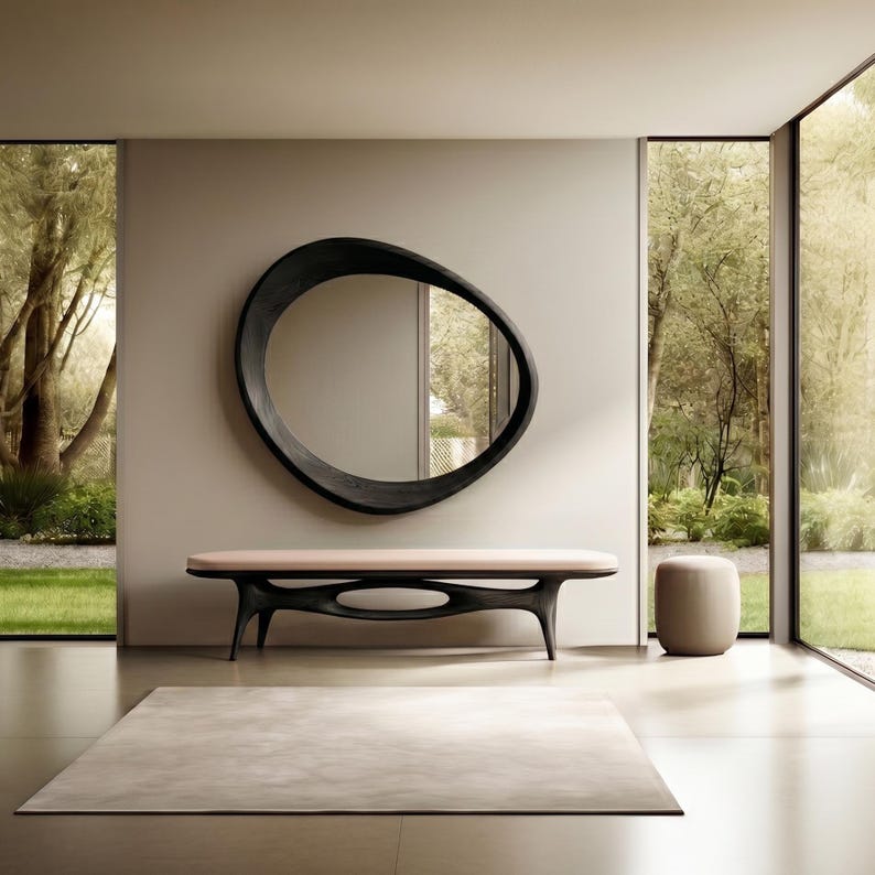 May include: A black, organic-shaped wall mirror with a thick frame hangs above a long, low bench. The bench features a black, curved base with a central cutout and a light pink, flat-topped seat. A small, cylindrical, light-colored stool sits nearby.