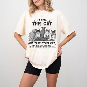 May include: Cream-colored t-shirt featuring a graphic of several cats in various poses. The text "ALL I NEED IS THIS CAT" is above the cat illustration, with additional text below. The shirt has short sleeves.