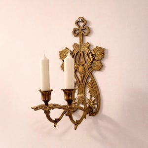 May include: Ornate gold-colored wall sconce with two candle holders. The sconce features a decorative design with floral and leaf motifs, and two white candles. The sconce is mounted on a light-colored wall.