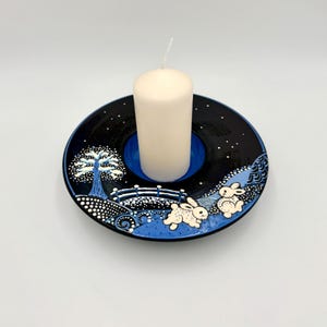 Può includere: Un portacandele in ceramica con un design a scena invernale. Il supporto è nero con un interno blu e presenta una candela bianca. Il design include un albero, un ponte e due conigli.