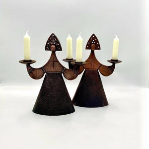 May include: Two decorative candle holders in a unique, stylized design. Each holder is made of dark brown metal and shaped like a figure with a conical body and a head with a decorative pattern. Each figure holds a round plate with a white candle.