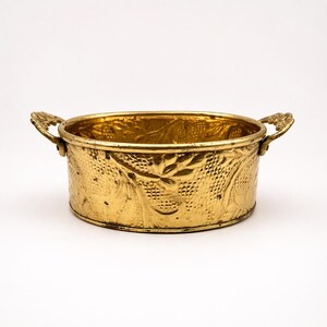 Könnte beinhalten: Ein goldfarbener, oval geformter Metallbehälter mit zwei verzierten Griffen. Die Außenseite des Behälters weist ein strukturiertes Muster mit geprägten Blumen- und Blattmotiven auf. Der Innenraum ist ebenfalls goldfarben, und das Stück ist vor einem weißen Hintergrund platziert.
