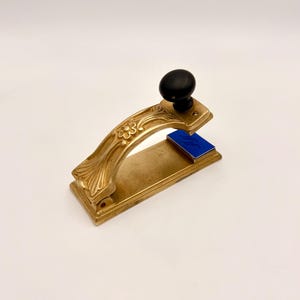 Op de afbeelding: Een vintage messing bureaustempel met een zwarte knop en een blauw stempelkussen. De stempel heeft een sierlijk bloemmotief op de handgreep. De stempel wordt waarschijnlijk gebruikt voor officiële documenten of persoonlijke correspondentie.