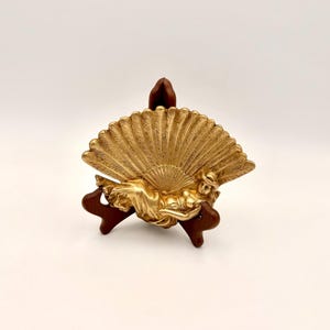 May include: A gold-colored decorative fan with a reclining figure, set on a dark wooden stand. The fan has a detailed feather design. The figure is in a relaxed pose, adding an artistic touch to the piece.