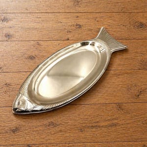 Könnte beinhalten: Eine silberfarbene, fischförmige Servierplatte. Die Platte hat einen detaillierten Fischkopf und Schwanz, mit einem ovalen Servierbereich. Das glänzende Metalltablett liegt auf einer Holzoberfläche.