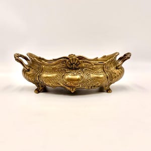 May include: An elongated, metallic gold-toned bowl featuring ornate, raised relief patterns across its exterior. The vessel includes two curved handles on opposite ends, a scalloped rim, and is supported by four small, decorative feet.