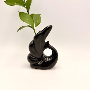 May include: A glossy black ceramic vase with a unique, sculptural design. The vase features a circular opening and a curved, organic shape. Green leafy plant stems are inserted into the vase, creating a striking contrast against the black surface.