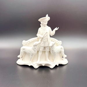 Puede incluir: Figurilla de color marfil que representa a un hombre con atuendo del siglo XVIII, sosteniendo una fusta, flanqueado por dos perros. El hombre lleva sombrero y abrigo. La base tiene un diseño decorativo y curvo.