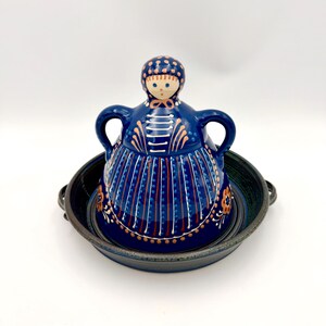 May include: A blue ceramic serving dish with a lid shaped like a doll. The dish has a round base and two handles. The lid features a painted face and a patterned dress with orange and white accents. The dish is on a white background.